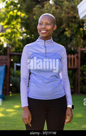 Afroamerikanerin, die im sonnendurchfluteten Garten in der Nähe des Spielsets steht und sportliches Top und Smartwatch trägt. Outdoor, Garten, sonnig, Fitness, Wellness, aktiv Stockfoto