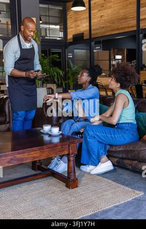 Männlicher Kellner, der die Bestellung von zwei Frauen auf einem Ledersofa im Café mit Tablet und Kaffeetassen entgegennimmt. Barista, Gastfreundschaft, lässig, Lounge, Denim, Anhänger, Pflanzen Stockfoto