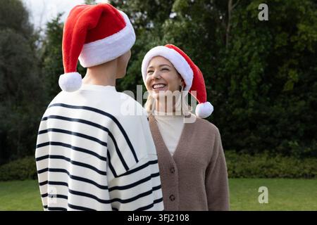 Paare stehen auf Grasrasen mit Weihnachtsmützen, lächelnde Frau, Mann mit gestreiftem Pullover. Duo, Outdoor, festlich, Urlaub, Strickjacke, kopfbedeckung, Eve Stockfoto