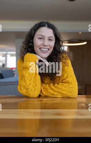 Frau Mitte 20 lehnte sich auf polierten Holztisch in der Lounge und lächelte in senfgelben Pullover. Wohnzimmer, gemütlich, Strickwaren, Anhänger, Innenausstattung, Lebensstil Stockfoto