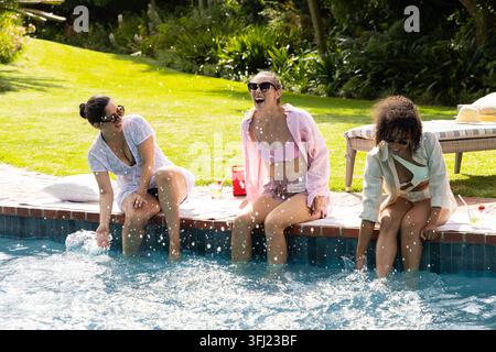 Verschiedene Freundinnen, die am Rand des Heimschwimmbeckens sitzen, leichte Überwürfe tragen und Wasser spritzen. Hinterhof, sonnig, draußen, Bi Stockfoto