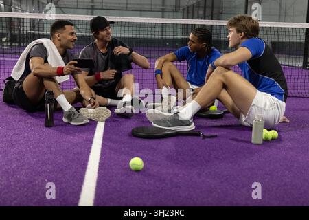 Verschiedene männliche Mannschaftskameraden sitzen auf dem purpurpurfarbenen Platz mit sportlicher Ausrüstung, die die Paddles und das Smartphone halten. Teamwork, Schläger, Bälle, Flaschen, entspannt, Arena Stockfoto