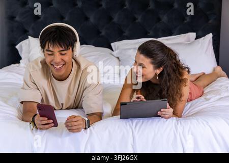 Verschiedene Paare liegen im Schlafzimmer, tragen legere Kleidung, verwenden Smartphone und Tablet. Gemütlich, entspannt, intim, Kopfteil, Kopfhörer, Smartwatc Stockfoto