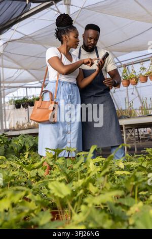 Kollegen im Gewächshaus unter Topfpflanzen, die auf das Telefon zeigen, einen Bräunungsbeutel tragen, eine dunkle Schürze tragen. Gärtnerei, Teamarbeit, Gartenbau, Einzelhandel, consulta Stockfoto