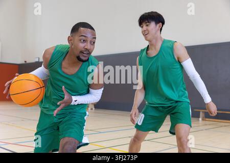 Verschiedene männliche Teamkollegen dribbeln orangen Basketball und bewegen sich auf dem Fitnessplatz in grünen Trikots. Athleten, Ball, Hartholz, Bank, Training, Teamwork, Moti Stockfoto