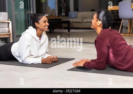 Verschiedene Freundinnen liegen auf Yogamatten zu Hause, tragen einen weißen Hoodie und einen kastanienbraunen Pullover, Telefon in der Nähe. Fitness, Partnerschaft, Konver Stockfoto