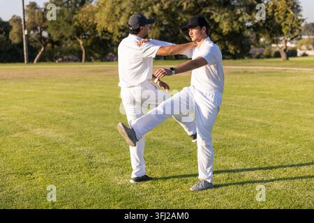 Verschiedene männliche Spieler wärmen sich auf, dehnen sich auf dem Parkfeld in weißem Kit, dunkle Kappen, Handgelenkstracker. Athleten, Cricket, Outdoor, Rasen, Uniformen, duo, Su Stockfoto