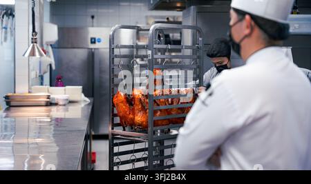 zwei Köche bereiten Enten zum Kochen vor, professioneller männlicher Koch in einer weißen Uniform beobachtet das Fleischbraten in einem feurigen Rotisserie-Ofen in einer kommerziellen Küche Stockfoto