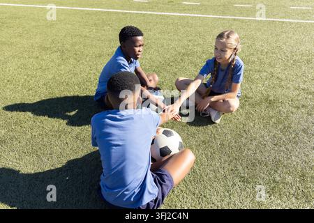 Verschiedene Kinder sitzen im Kreis auf dem Rasen, halten Fußball und stapeln Hände in blauen Hemden. Trio, Feld, Meeting, draußen, Sonnenlicht, Teamarbeit, Stockfoto