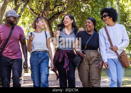 Verschiedene Freunde, die auf schattigen Parkwegen laufen, halten Kaffeetassen und Telefone, tragen Sonnenschirme und Taschen. Gruppe, Begleiter, Outdoor, Urban, sonnenbeleuchtet, Schutzdach, Stockfoto