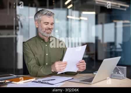 Geschäftsmann, der Dokumente und Finanzanalysen am Büroarbeitsplatz mit Laptop überprüft. Professioneller Mann arbeitet an Unternehmensstrategie und Projektplanung, während er wichtige Geschäftspapiere liest. Stockfoto