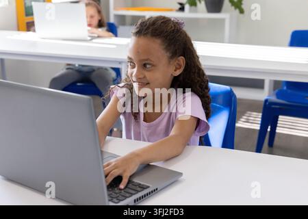 Weibliche Klassenkameraden sitzen an langen weißen Tischen, Mädchen in lila T-Shirt, die auf silbernem Laptop tippen. Schüler, Klassenzimmer, Computer, Lernen, Bildung Stockfoto