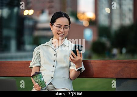 Junge asiatische Frau lächelt und benutzt ein Smartphone im Freien Stockfoto