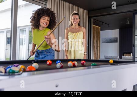 Verschiedene Freundinnen in grüner Decke lehnen sich zu Hause über den Billardtisch und halten einen Queuestift. Billard, Duo, spielen, Lounge, Tageslicht, moderne fenster Stockfoto