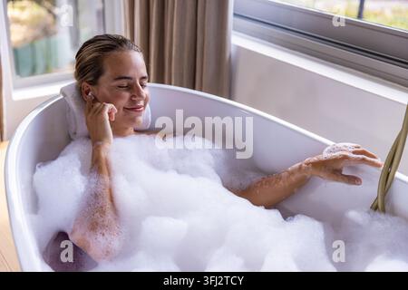 Frau, die sich in einer freistehenden Wanne mit dickem Schaum im Badezimmer zurücklehnt und Ohrstöpsel nahe gerolltem Handtuch berührt. Bad, Blasen, Tageslicht, Fenster, Grün, ruhig, Stockfoto