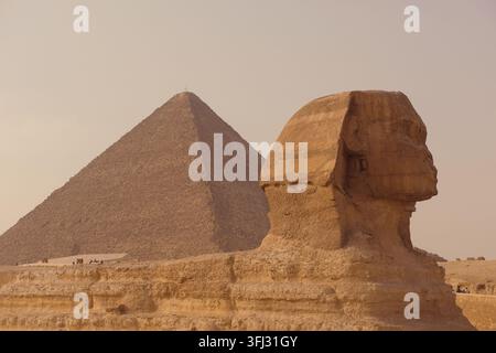 Das verwitterte Seitenprofil der Großen Sphinx steht vor der hoch aufragenden Großen Pyramide des Khufu in Gizeh unter einem dunstigen Wüstenhimmel. Zeitlos majestätisch Stockfoto