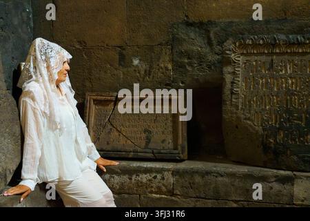 Armenien, 8.-15.2026: Frau in Weiß posiert neben gemeißelten Steintafeln und verwitterten Mauern in einer historischen religiösen Stätte, die kulturelle h Stockfoto