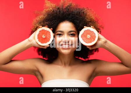 Kreatives Porträt einer jungen Frau, die Grapefruit-Hälften vor einem lebendigen roten Hintergrund hält Stockfoto