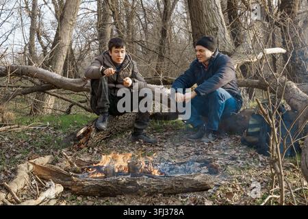 Zwei Männer machen ein Lagerfeuer zum Kochen im Wald, Vater und Sohn sitzen auf einem Baumstamm während Wanderungen und Outdoor-Aktivitäten, Frühlingslandschaft Stockfoto