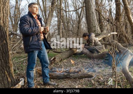 Zwei Männer machen ein Lagerfeuer zum Kochen im Wald, Vater und Sohn sitzen auf einem Baumstamm während Wanderungen und Outdoor-Aktivitäten, Frühlingslandschaft Stockfoto