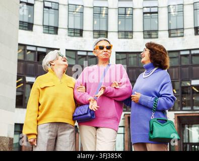 Drei fröhliche, ältere Freundinnen in hellen Pullovern und Sonnenbrillen gehen gemeinsam in der Nähe eines modernen Gebäudes. Lifestyle- und Menschenkonzept. Stockfoto