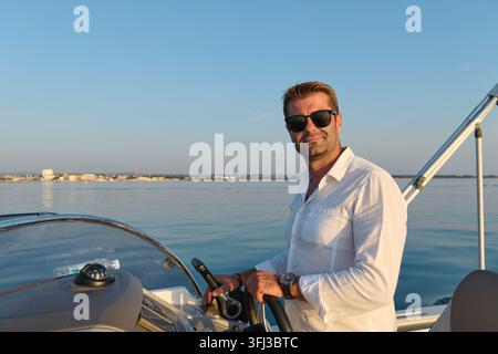 Ein entspannter Mann in einem weißen Leinenhemd und Sonnenbrille steuert ein Motorboot vor der Küste von Umag zur goldenen Stunde, ruhige Adriawasser und ferne Küste e Stockfoto