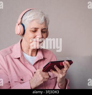 Eine glückliche ältere Frau, die ihre Kopfhörer und ihr Mobiltelefon benutzt, um einen Podcast zu hören, während sie zu Hause entspannt. Stockfoto