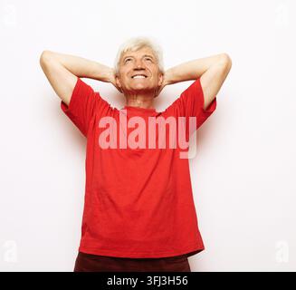 Attraktiver Senior trägt ein rotes T-Shirt, entspannt und streckt sich mit Armen und Händen hinter Kopf und Nacken, lächelt glücklich. Stockfoto