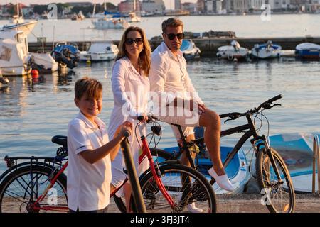 Eine Familie mit drei Eltern und ihrem Sohn genießt eine entspannte Radtour entlang eines Hafenbeckens zur Goldenen Stunde, mit vertäuten Booten und warmem Sonnenlicht Stockfoto
