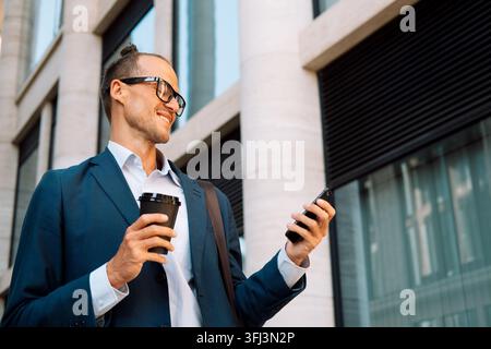 Junger Mann, lässig gekleidet, hält eine Tasse Kaffee zum Mitnehmen, lächelt glücklich und nutzt sein Smartphone in der Stadt. Stockfoto