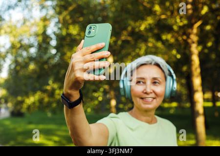 Zufriedene und lächelnde 60-jährige asiatische Frau mit Kopfhörern telefoniert mit ihrem Enkelkind per Videoanruf, während sie einen Spaziergang im grünen Park genießt. Hobby und Freizeit Stockfoto