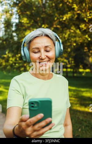 Zufriedene und lächelnde 60-jährige asiatische Frau mit Kopfhörern telefoniert mit ihrem Enkelkind per Videoanruf, während sie einen Spaziergang im grünen Park genießt. Hobby und Freizeit Stockfoto