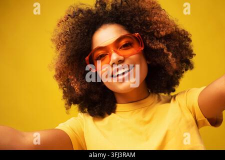 Junge afroamerikanische Bloggerin macht Selfie, posiert lächelnd zur Kamera im Studio vor gelbem Hintergrund, trägt gelbes T-Shirt und orangefarbene Sonnenbrille Stockfoto