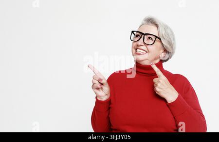 Lächelnde ältere grauhaarige Frau, isoliert auf grauem Studiohintergrund, zeigt mit dem Finger auf leeren Kopierraum daneben Stockfoto