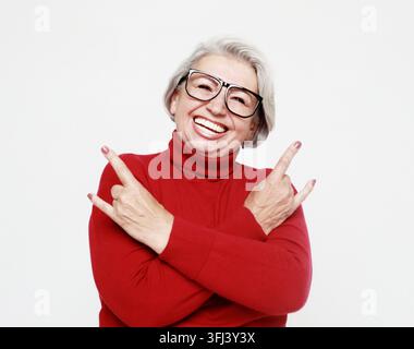 Porträt einer älteren grauhaarigen Frau, die einen roten Pullover und eine Brille trägt und über weißem Hintergrund ein Rock-and-Roll-Symbol macht Stockfoto
