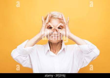 Seniorin mit grauen Haaren trägt ein weißes Hemd und macht eine OK-Geste in der Nähe der Augen Stockfoto