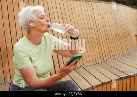 Eine ältere, glückliche Rentnerin sitzt nach einem Lauf auf einer Holzbank und trinkt Wasser. Stockfoto