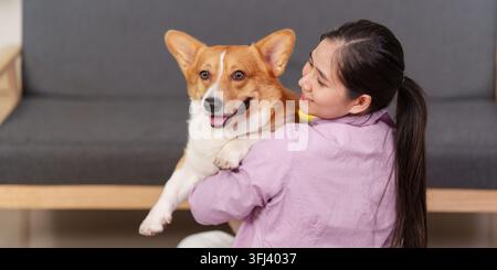 Eine asiatische Frau lächelt, während sie einen glücklichen Pembroke Welsh Corgi in ihren Armen hält, vor einem grauen Sofa. Stockfoto