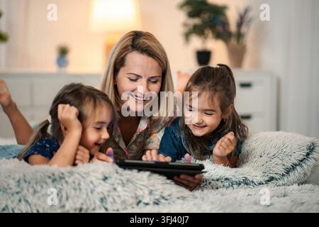 Glückliche Mutter und Töchter, die im Bett zusammen an einem Digitaltablett sitzen Stockfoto