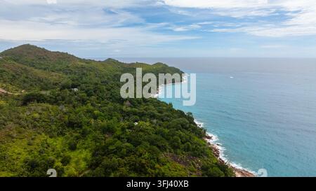 Eine ruhige Luftaufnahme von dicht bewaldeten Hügeln, die sich zu einer felsigen Küste hinabziehen und auf das ruhige, türkisfarbene Wasser treffen. Praslin, Seychellen. Stockfoto