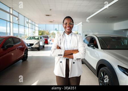 Junge erwachsene Geschäftsfrau lächelt, steht mit verschränkten Armen in einem Autohaus Stockfoto