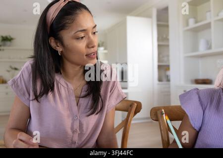 Afroamerikanische Mutter in Bluse, die sich an einen hölzernen Küchentisch lehnt, und ihre Tochter, die am Tisch sitzt und einen Bleistift benutzt. Erwachsene, Kind, Zuhause, Esszimmer, Stockfoto