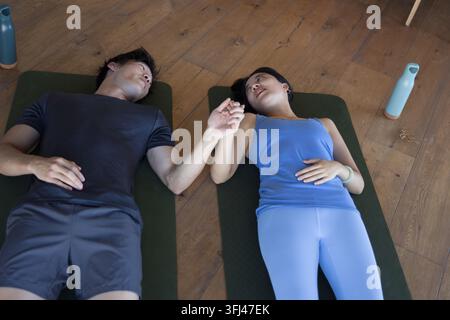 Asiatisches Paar liegt auf Yogamatten auf Dielenboden in Sportkleidung, hält sich an den Händen, Wasserflaschen in der Nähe. Zusammengehörigkeit, Balance, Wellness, Achtsamkeit, Ruhe Stockfoto