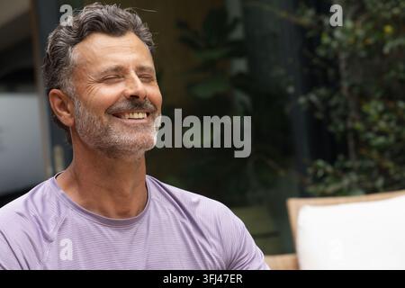 Mittelalter Mann lächelt mit geschlossenen Augen, sitzt auf Terrassenstuhl und trägt lila Sporthemd. Außenbereich, reif, Gelassenheit, Porträt, Nahaufnahme, Terrasse Stockfoto