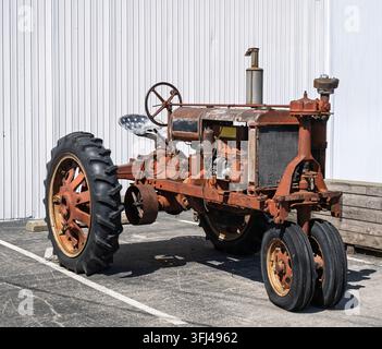 Rostiger Vintage-Traktor, der auf Beton geparkt ist. Große schwarze Reifen und Metallteile zeigen Abnutzungsspuren. Weißer vertikaler Wandverkleidung bildet den Hintergrund. Sonnenlicht wirft Schatten Stockfoto