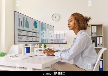 Finanzanalyst, der Geschäftscharts auf Dual-Bildschirmen in einem hellen Büro überprüft Stockfoto