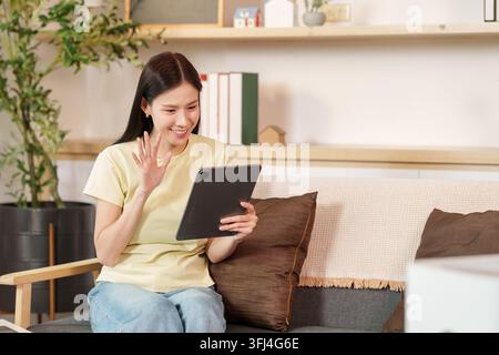 Lächelnde junge asiatische Frau, die auf einem bequemen Wohnzimmersofa sitzt und einen Videokonferenz-Anruf auf ihrem Tablet-Gerät macht. Stockfoto