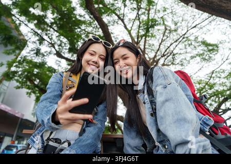 Niedrigwinkelaufnahme von zwei lächelnden asiatischen Touristinnen, die im Freien in der Stadt auf ein Smartphone hinunterschauen. Stockfoto