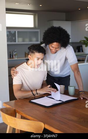 Verschiedene männliche Freunde, die sich über den Küchentisch lehnen und Papiere in einem schwarzen Portfolio mit Tassen überprüfen. Zusammenarbeit, Dokumente, Planung, Arbeitsbereich, Homeoffice Stockfoto