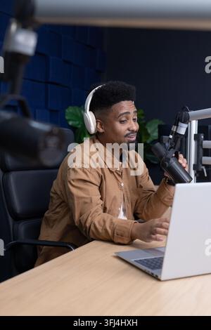 Erwachener afroamerikanischer Mann mit weißen Kopfhörern, der in ein Mikrofon an einem Boom spricht und einen Laptop im Studio verwendet Stockfoto